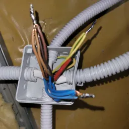 Szara puszka instalacyjna SIMET NS5 z podłączonymi przewodami elektrycznymi o różnych kolorach, zabezpieczonymi końcówkami tulejkowymi, wychodzącymi z karbowanych peszli elektrycznych ELEKTRO.