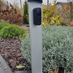 Szara kolumna z czarnym czytnikiem zbliżeniowym zamontowana w ogrodzie, widoczne rośliny i kostka brukowa, na kolumnie krople deszczu.