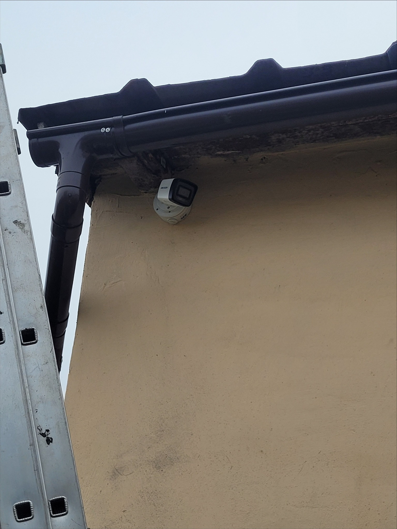 Kamera monitoringu zamontowana pod rynną na elewacji budynku, widoczna drabina oparta o ścianę.