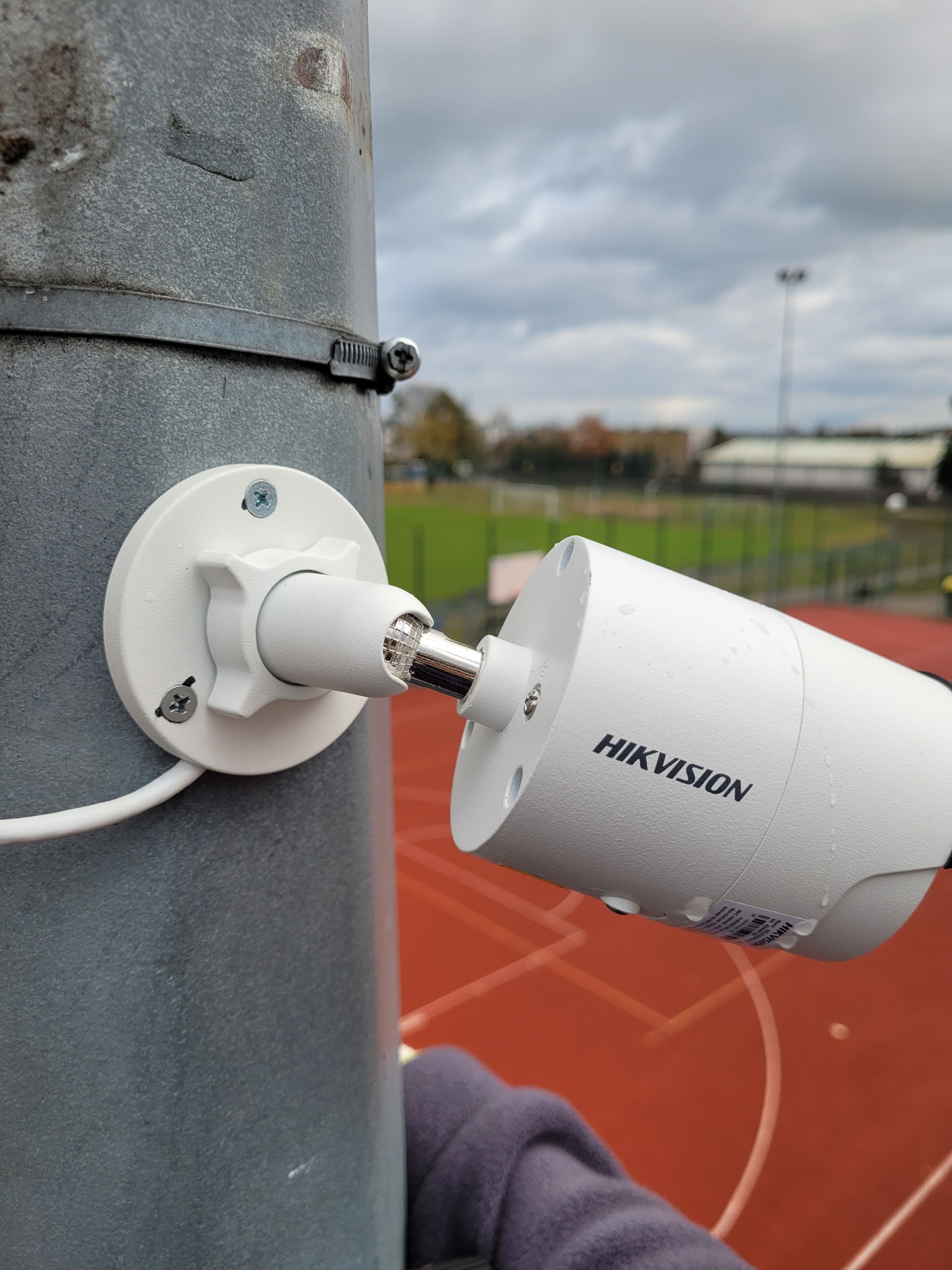 Biała kamera monitoringu marki Hikvision zamontowana na szarym słupie za pomocą metalowej obejmy i białego uchwytu, widok na boisko sportowe w tle.
