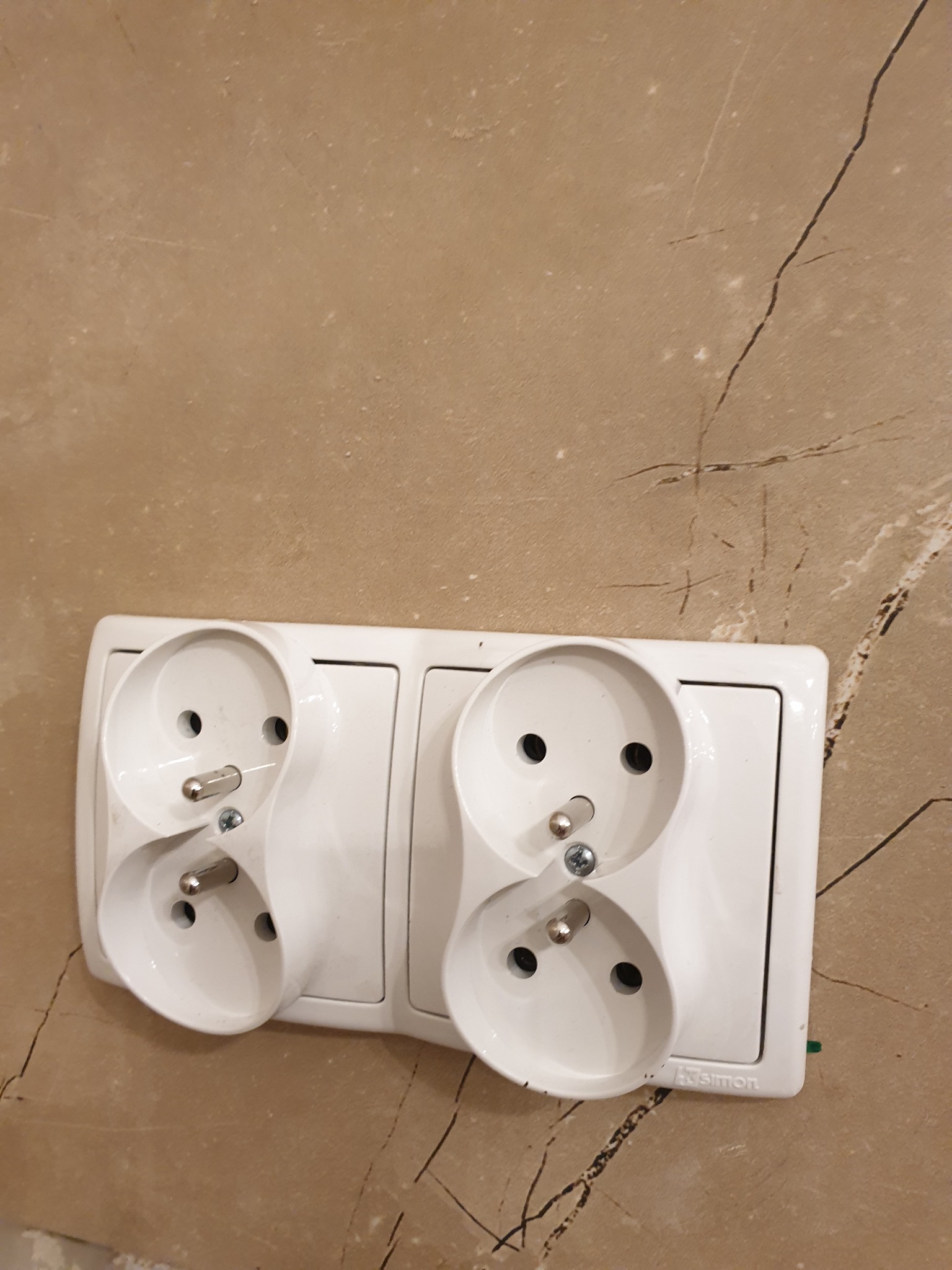 Podwójne, białe gniazdko elektryczne z uziemieniem, zamontowane na ścianie wykończonej płytkami imitującymi marmur. Widoczny wtyk w jednym z gniazd.