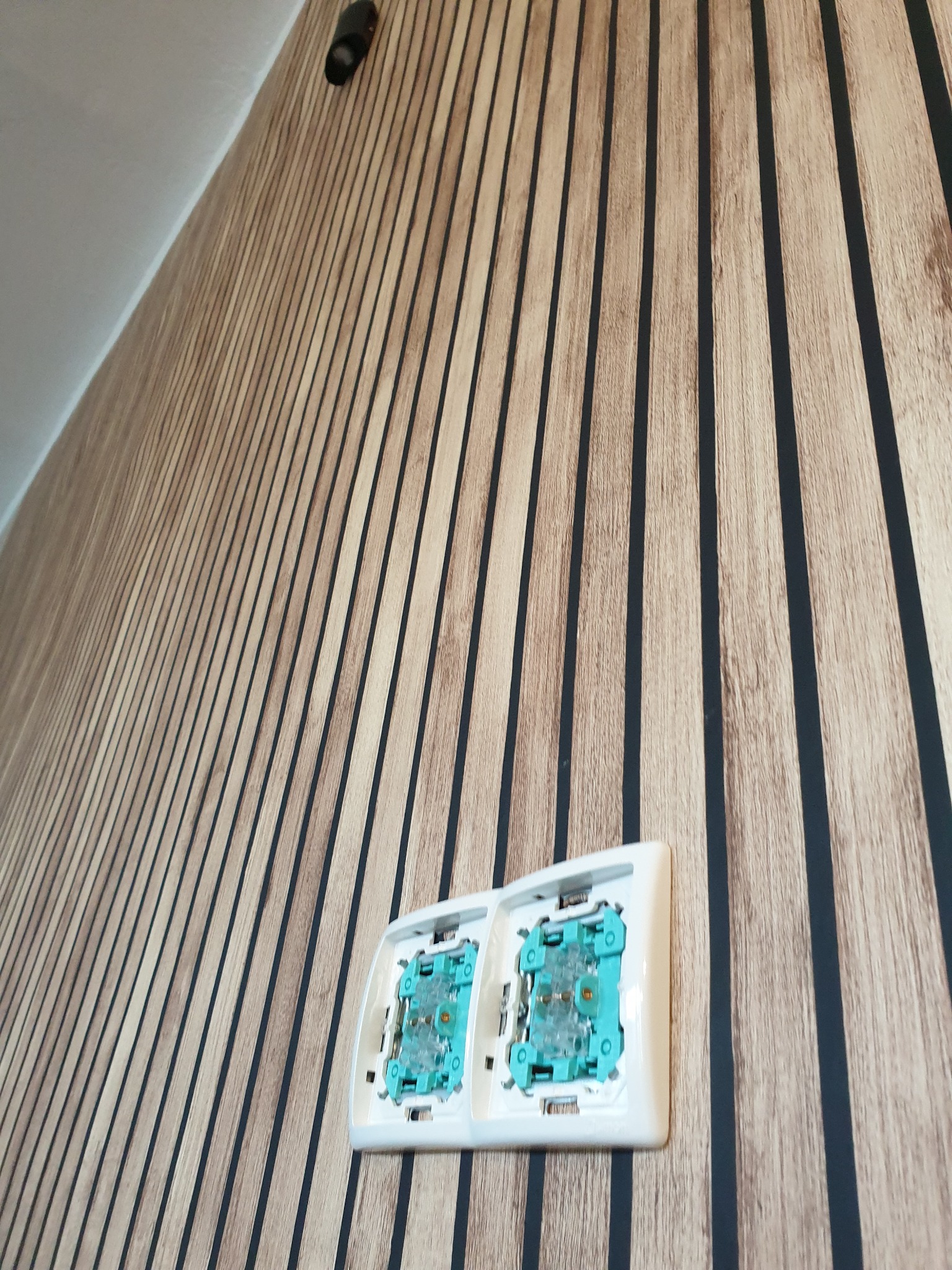 Dwa gniazdka elektryczne bez osprzętu, zamontowane na ścianie z dekoracyjną okładziną w pionowe pasy w odcieniach brązu i czerni; widoczny również czujnik ruchu pod sufitem.