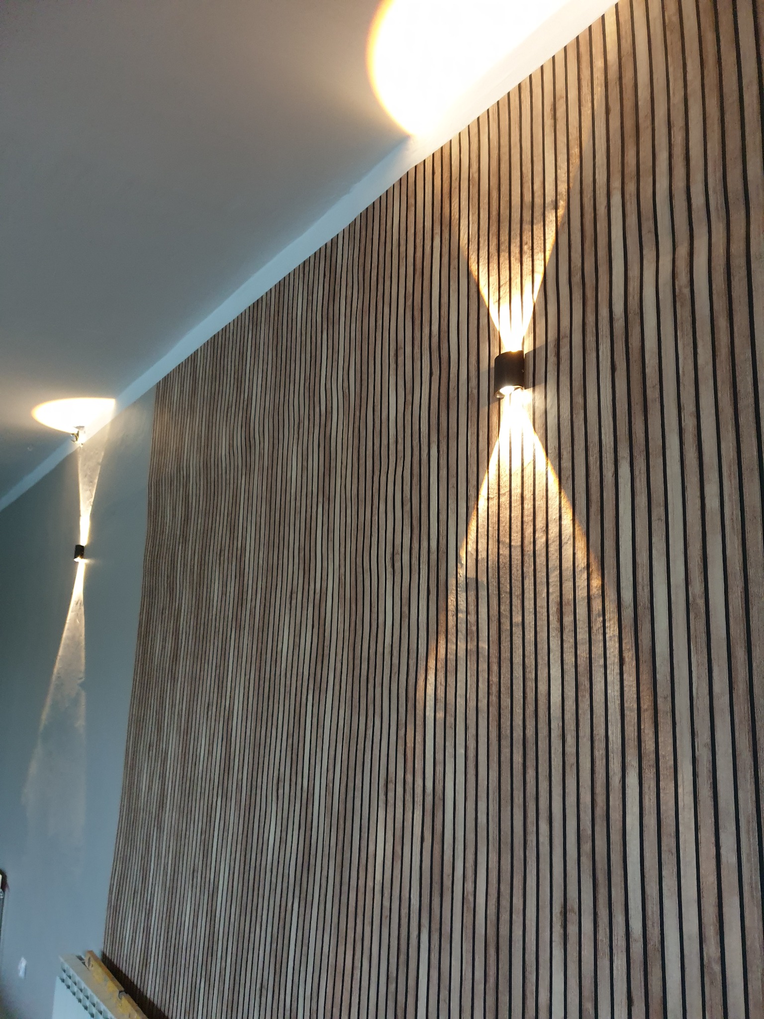 Dwa nowoczesne kinkiety ścienne oświetlające panel z pionowych drewnianych listew na ścianie.