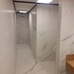 Wąskie wnętrze łazienki wykończone białymi płytkami imitującymi marmur z szarymi żyłkami, z widocznym fragmentem drewnianej umywalki i minimalistycznym oświetleniem sufitowym.