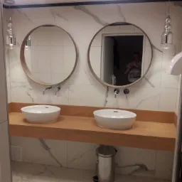 Nowoczesna łazienka z dwoma okrągłymi lustrami, białymi umywalkami nablatowymi na drewnianym blacie, marmurowymi płytkami na ścianach i podłodze oraz srebrnym koszem na śmieci.