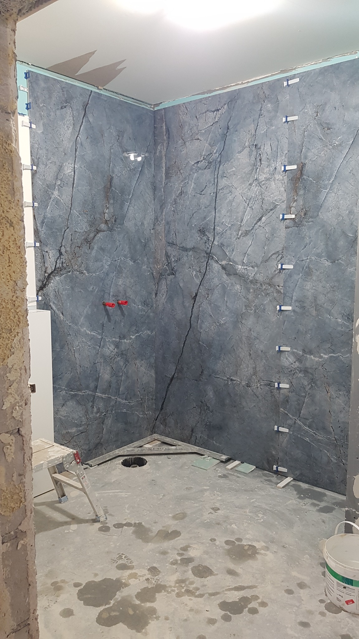 Montaż płytek imitujących marmur w kabinie prysznicowej, widoczne poziomice i plastikowe krzyżyki dystansowe, surowy beton na podłodze z plamami wody.