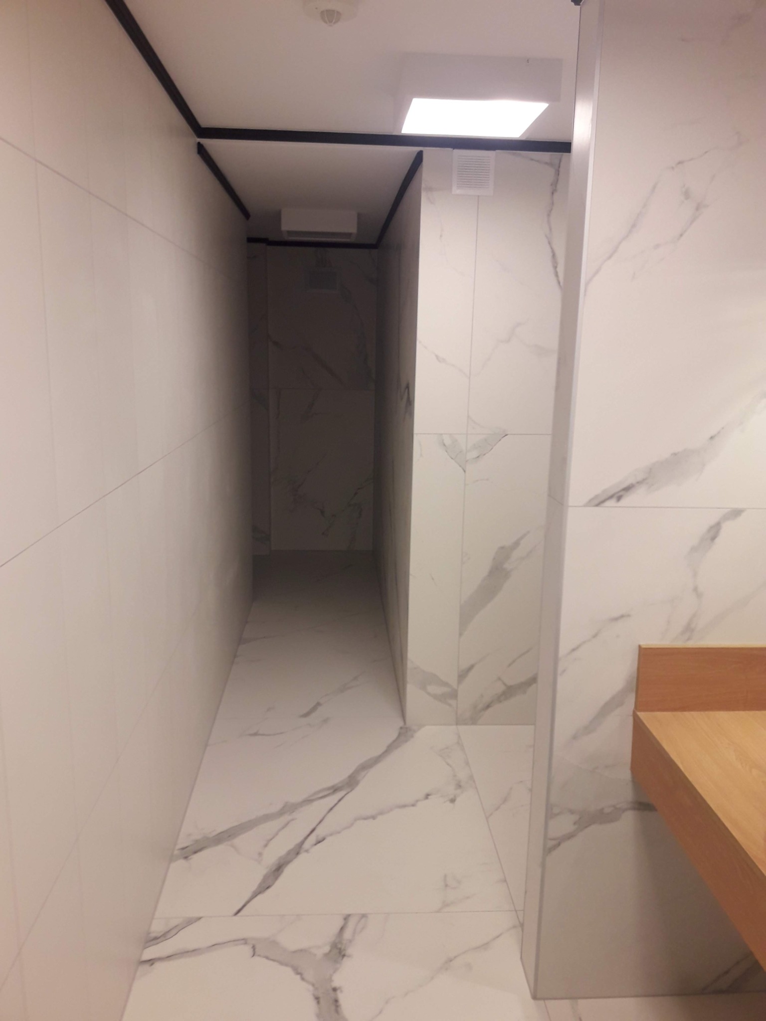 Wąskie pomieszczenie wyłożone białymi płytkami imitującymi marmur, z widoczną drewnianą półką po prawej stronie i czarną listwą sufitową.