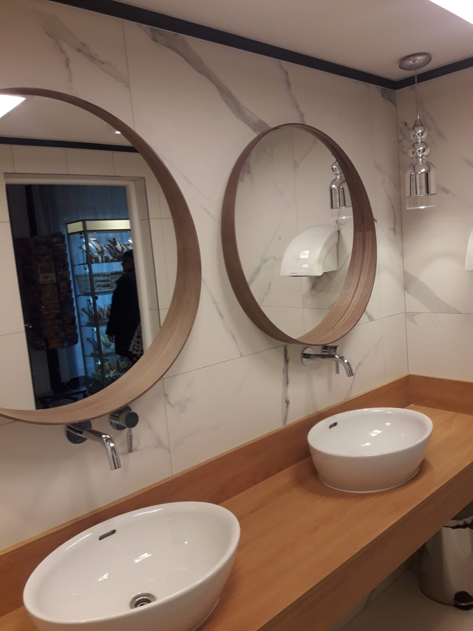 Dwie owalne umywalki nablatowe z drewnianymi ramami luster na tle marmurowej ściany i designerskich lamp sufitowych w nowoczesnej toalecie publicznej, z widocznym odbiciem sklepu w jednym z luster.