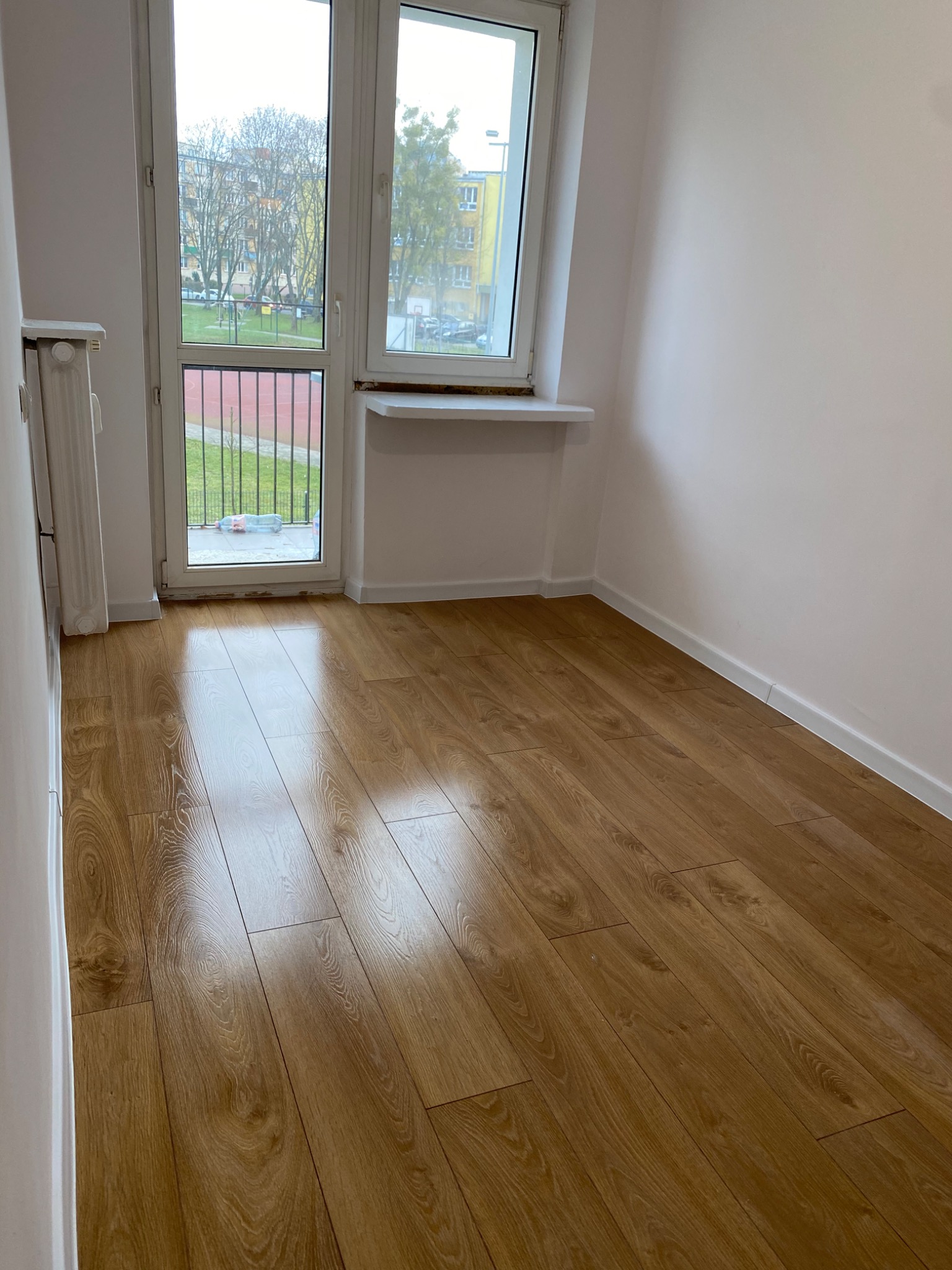 Jasne panele podłogowe z widoczną strukturą drewna, świeżo ułożone w pustym pokoju z białymi ścianami i oknem balkonowym z widokiem na plac zabaw.