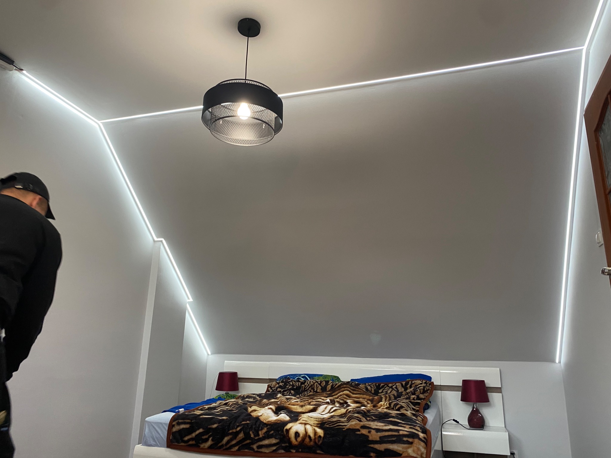 Sypialnia na poddaszu z ukośnymi ścianami oświetlonymi liniowym LED, z wiszącą lampą z ażurowym kloszem i łóżkiem przy ścianie.