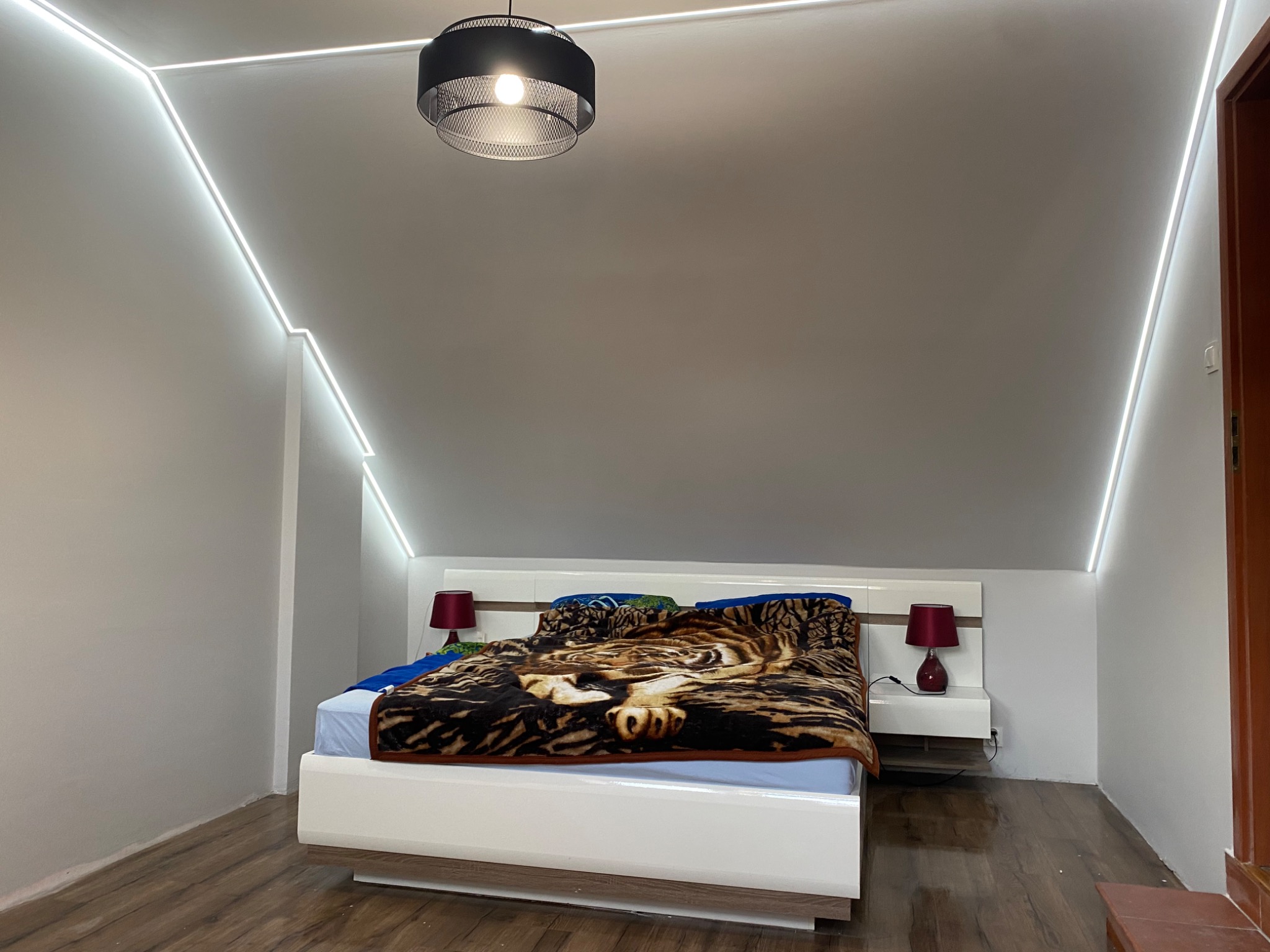 Pokój na poddaszu z drewnianą podłogą, białymi ścianami, oświetleniem LED wzdłuż skosów sufitu, łóżkiem z narzutą w tygrysi wzór i czarną lampą wiszącą.