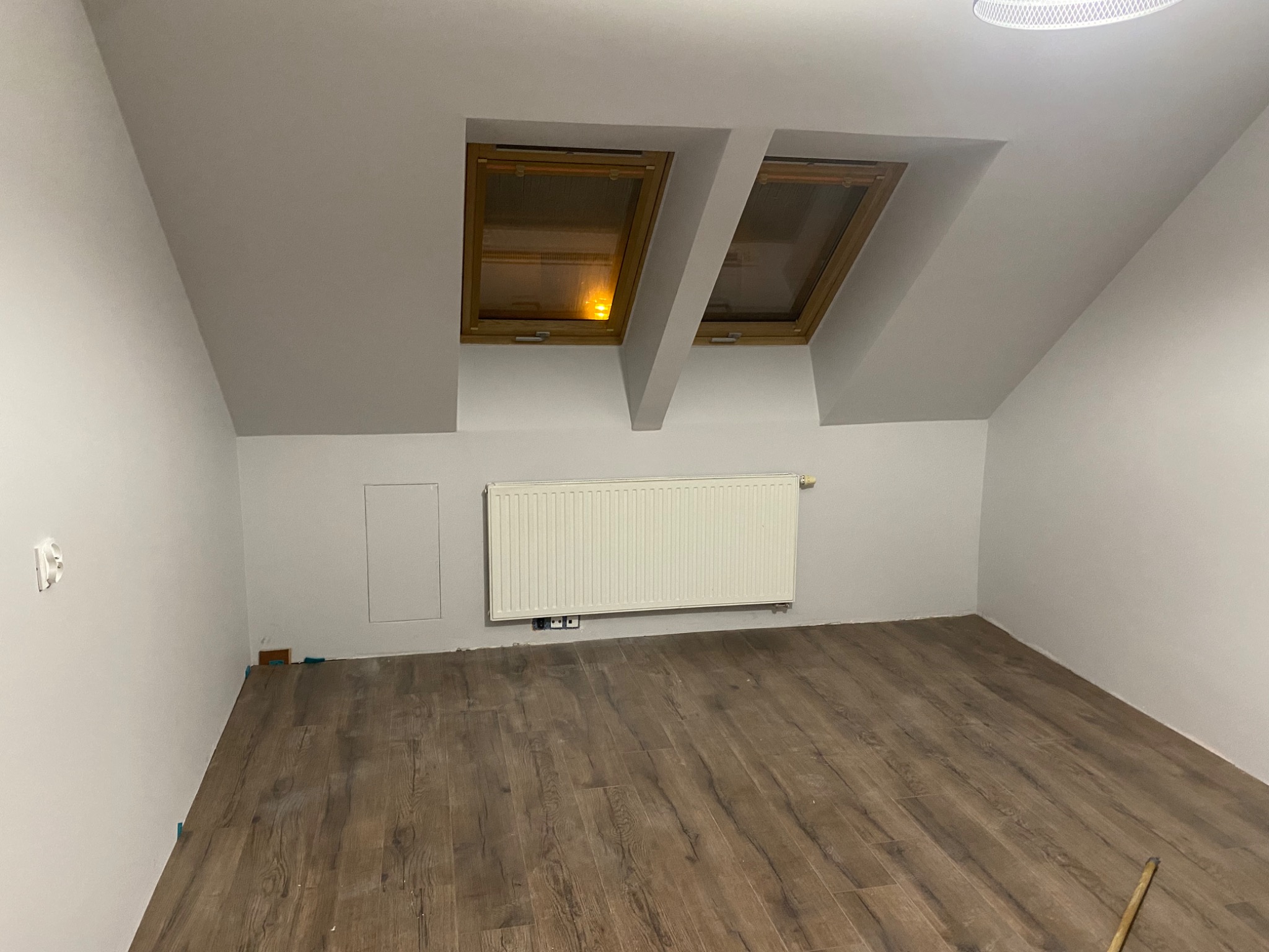 Poddasze z dwoma oknami dachowymi, świeżo ułożone panele podłogowe w odcieniach brązu, białe ściany i grzejnik. Widoczne niezapełnione szczeliny dylatacyjne przy ścianach.
