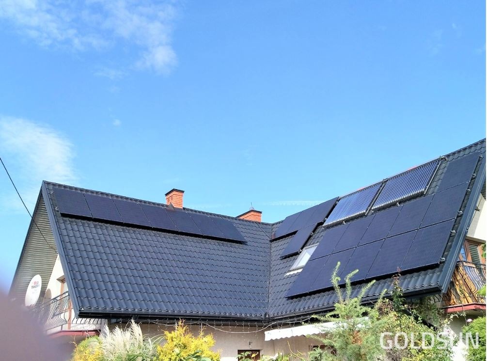 Czarny dach dwuspadowy z panelami fotowoltaicznymi i kolektorami słonecznymi, widok z dołu na tle błękitnego nieba.