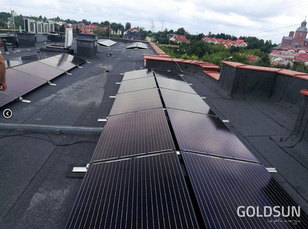 Panele słoneczne zamontowane rzędami na płaskim dachu budynku z widokiem na miasto i kościół w tle.