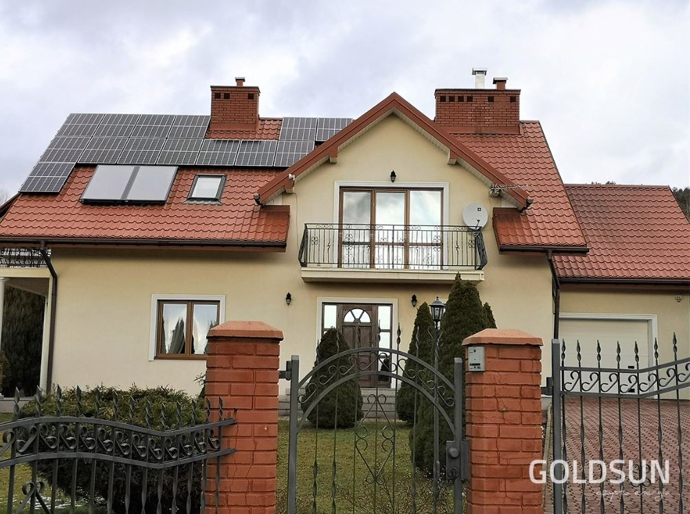 Dom jednorodzinny z panelami słonecznymi na dachu, widok zza ozdobnej, metalowej bramy z ceglanymi słupkami, logo firmy widoczne w prawym dolnym rogu.