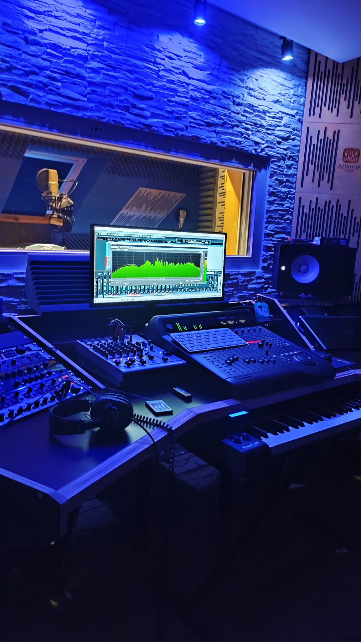 Profesjonalne stanowisko mikserskie z monitorem wyświetlającym parametry dźwięku, widok na kabinę nagrań z mikrofonem, oświetlone niebieskim światłem w pomieszczeniu wykończonym kamieniem...