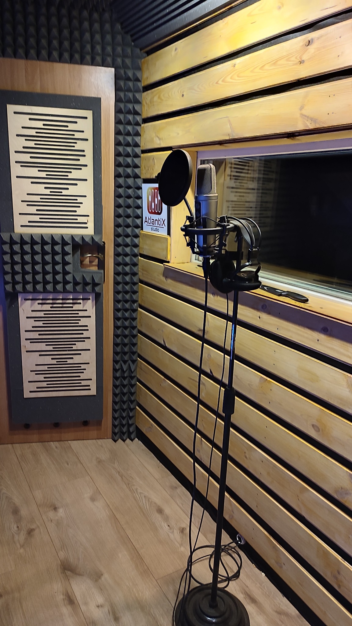 Wnętrze studia nagrań z mikrofonem na statywie, słuchawkami i panelami akustycznymi na ścianach, widoczne logo studia 'Atlantix'.