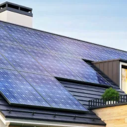 Panel słoneczny na dachu domu z oknem mansardowym, widok z dołu, błękitne niebo w tle.