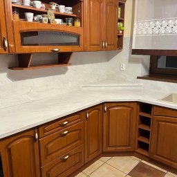 Mebelfach Mariusz Krupa - Klasyczna zabudowa kuchenna z drewnianymi frontami, jasnym blatem i przeszkloną szafką wiszącą, uzupełniona o dekoracyjne filiżanki na półce.