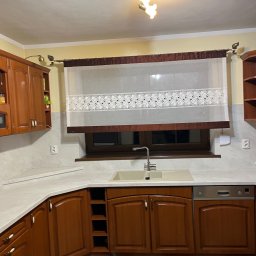 Mebelfach Mariusz Krupa - Klasyczna zabudowa kuchenna z drewnianymi frontami i jasnym blatem, Zawiercie. Widoczna armatura, okno z roletą i płytki podłogowe w szachownicę.