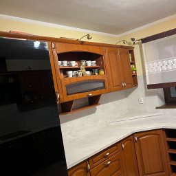 Mebelfach Mariusz Krupa - Drewniana zabudowa kuchenna z przeszklonymi szafkami wiszącymi, oświetleniem punktowym i białym blatem. Widoczny fragment czarnej lodówki i okno z firanką.