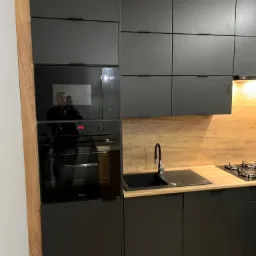 Nowoczesna, czarna zabudowa kuchenna z drewnianym blatem i okładziną ściany, z piekarnikiem Amica i odblaskiem osoby robiącej zdjęcie w szybie piekarnika i mikrofalówki.