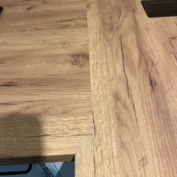 Fragment podłogi z paneli imitujących drewno, z widocznym czarnym gniazdkiem w podłodze i fragmentem blatu meblowego z widocznym napisem 'ATHLON EXTRUSIO'