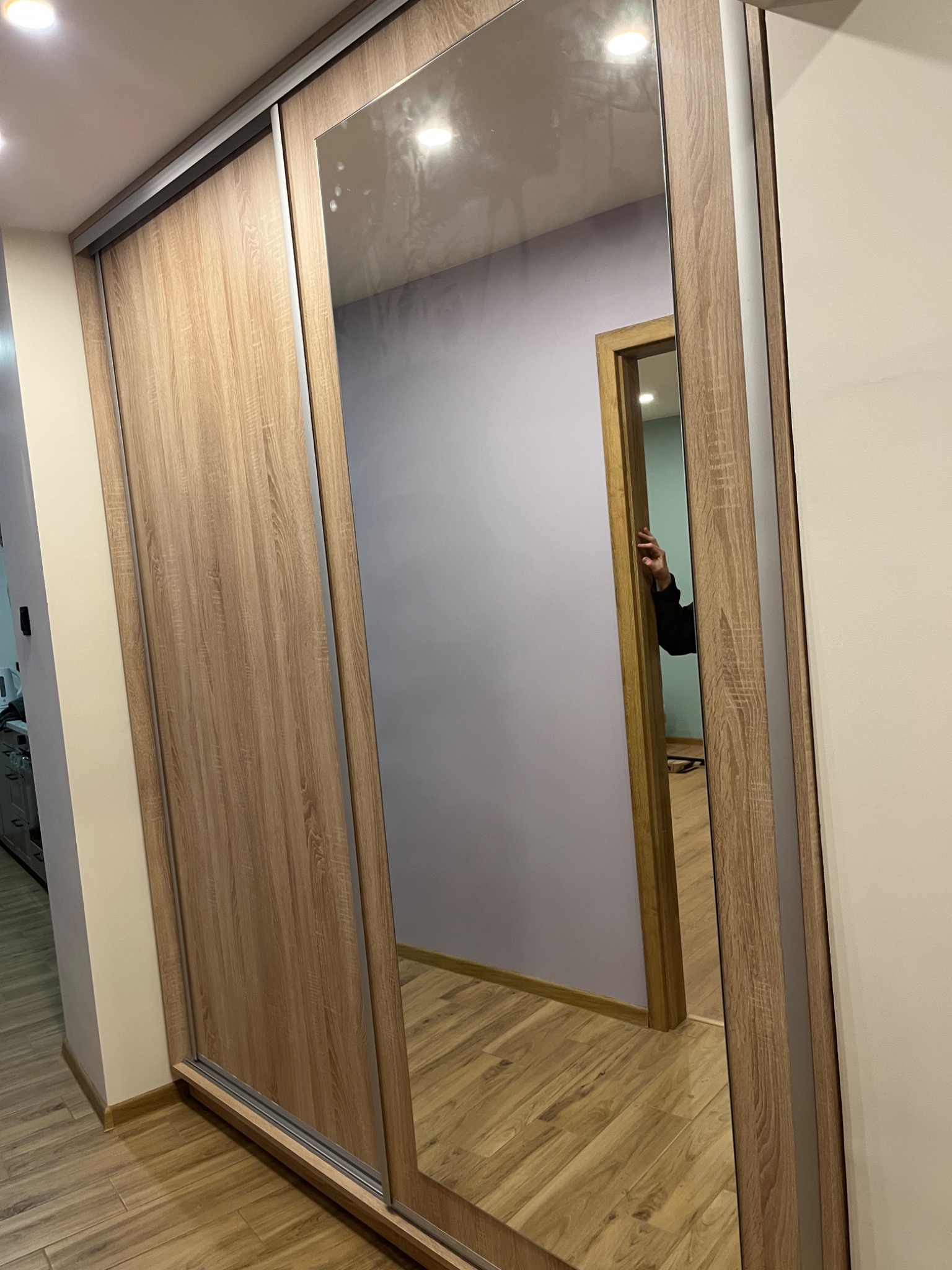 Szafa wnękowa z lustrem i przesuwnymi drzwiami o drewnianym wykończeniu, widoczna w korytarzu z parkietem. W lustrze odbija się fragment innego pomieszczenia.
