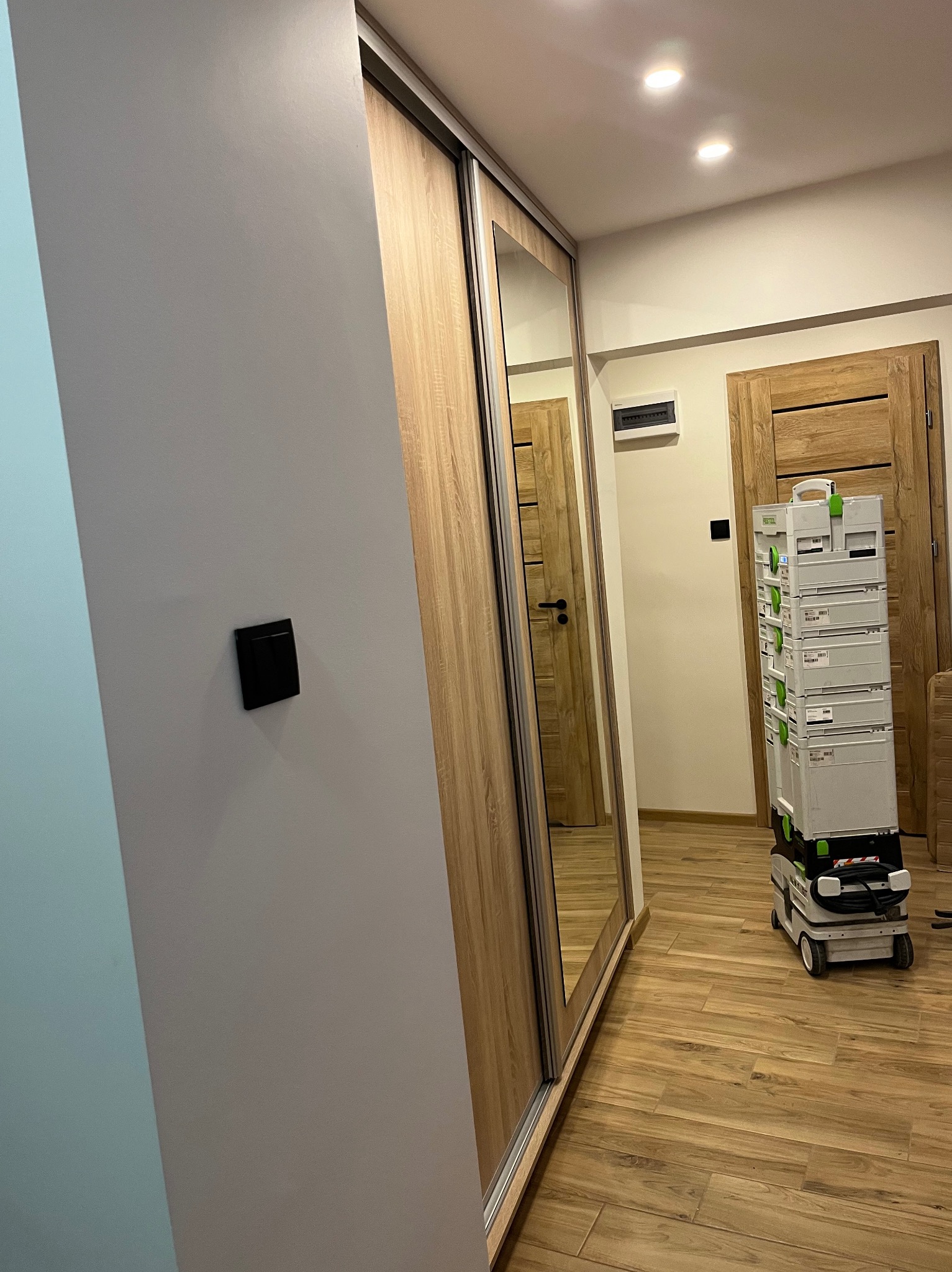 Nowoczesna szafa przesuwna z lustrem i drewnianym wykończeniem, widoczna na tle drewnianej podłogi i drzwi. Obok szafy stoi zestaw narzędzi.