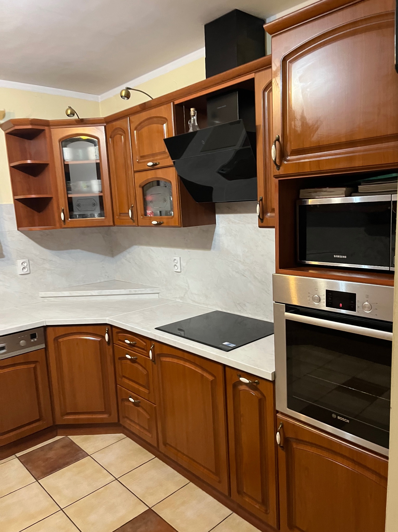 Klasyczna kuchnia z drewnianymi frontami, białym blatem i sprzętem AGD w zabudowie. Widoczny fragment podłogi z płytek w odcieniach beżu i brązu.