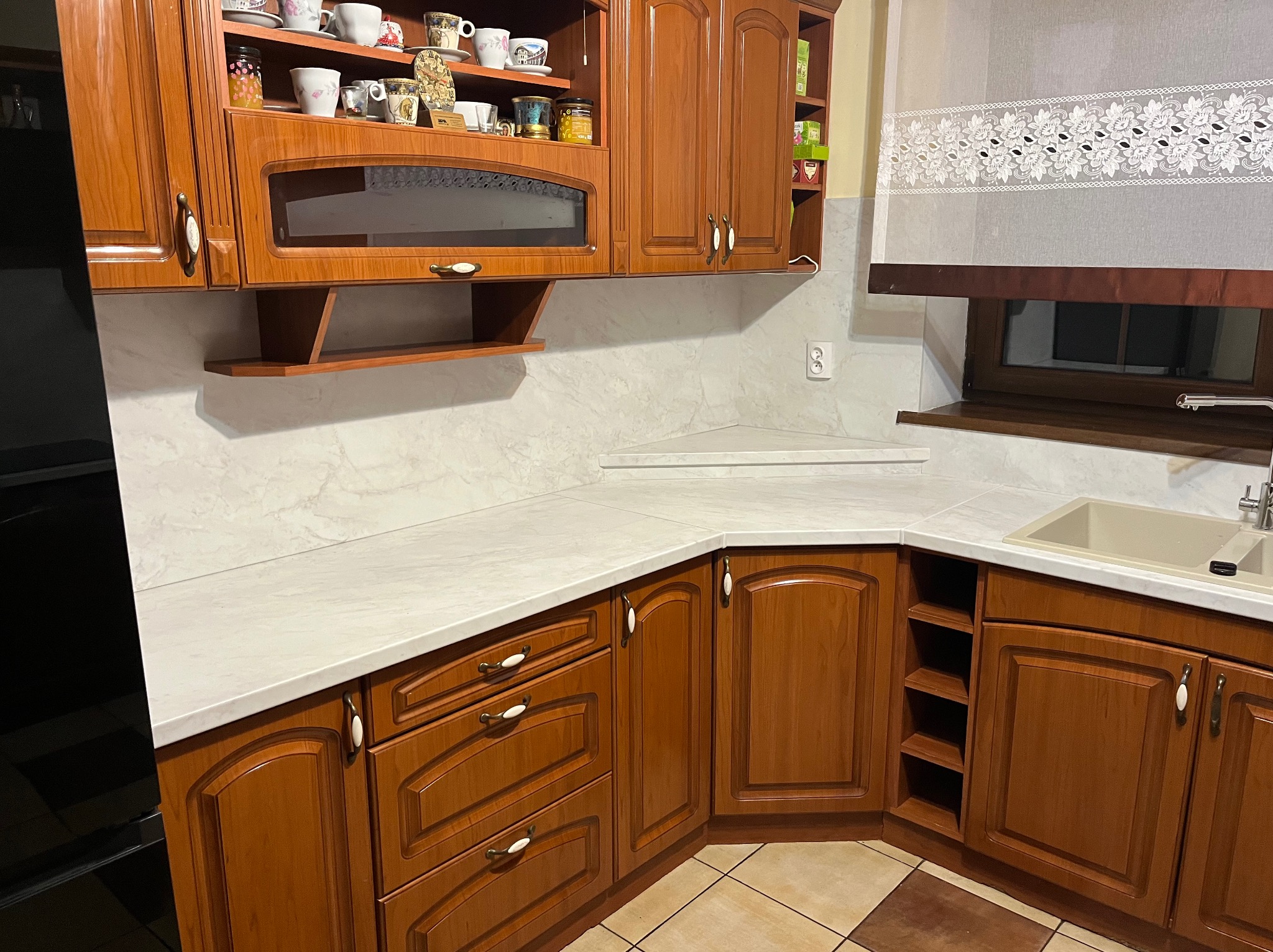 Klasyczna zabudowa kuchenna z drewnianymi frontami, jasnym blatem i przeszkloną szafką wiszącą, uzupełniona o dekoracyjne filiżanki na półce.