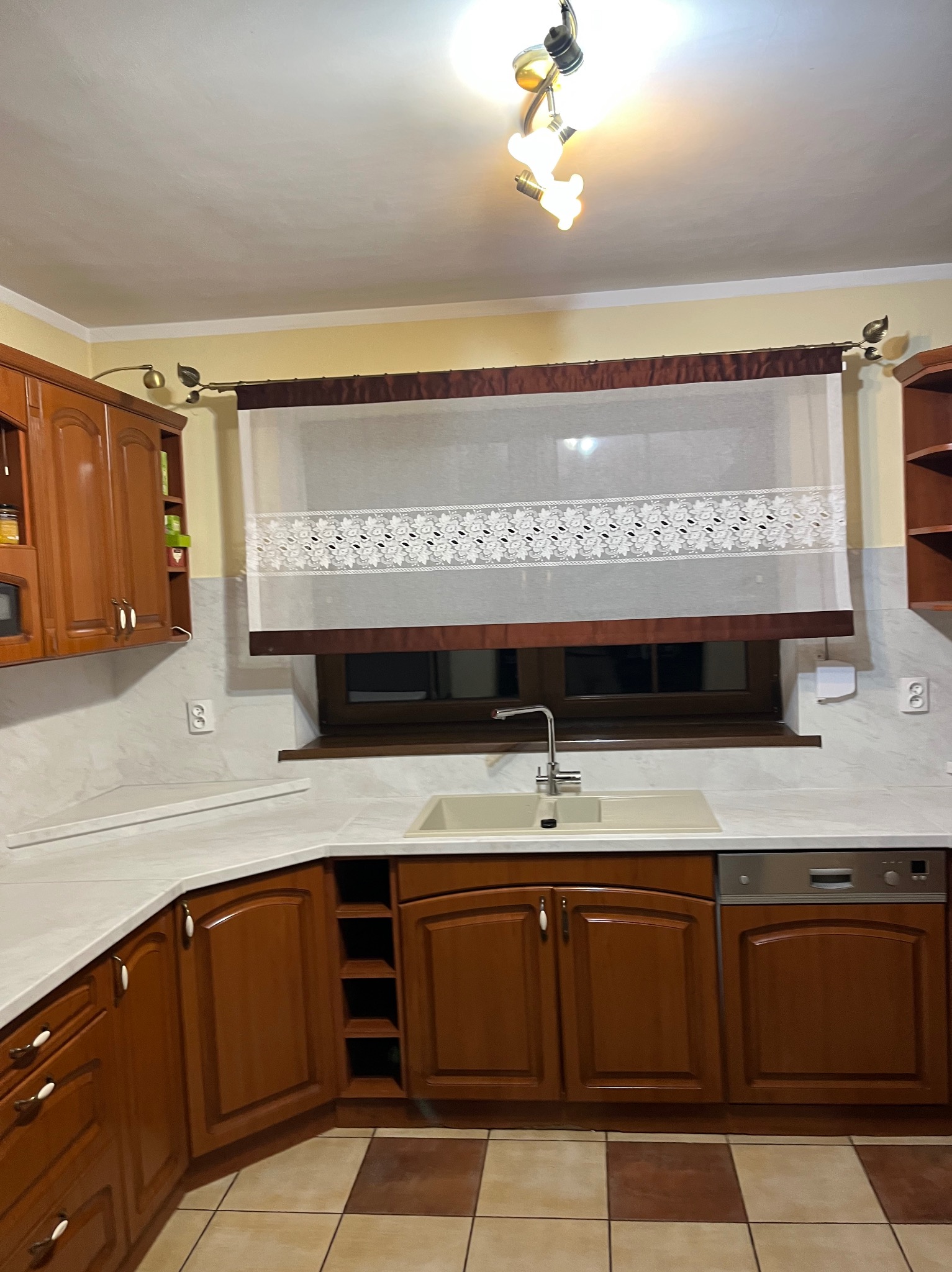 Klasyczna zabudowa kuchenna z drewnianymi frontami i jasnym blatem, Zawiercie. Widoczna armatura, okno z roletą i płytki podłogowe w szachownicę.