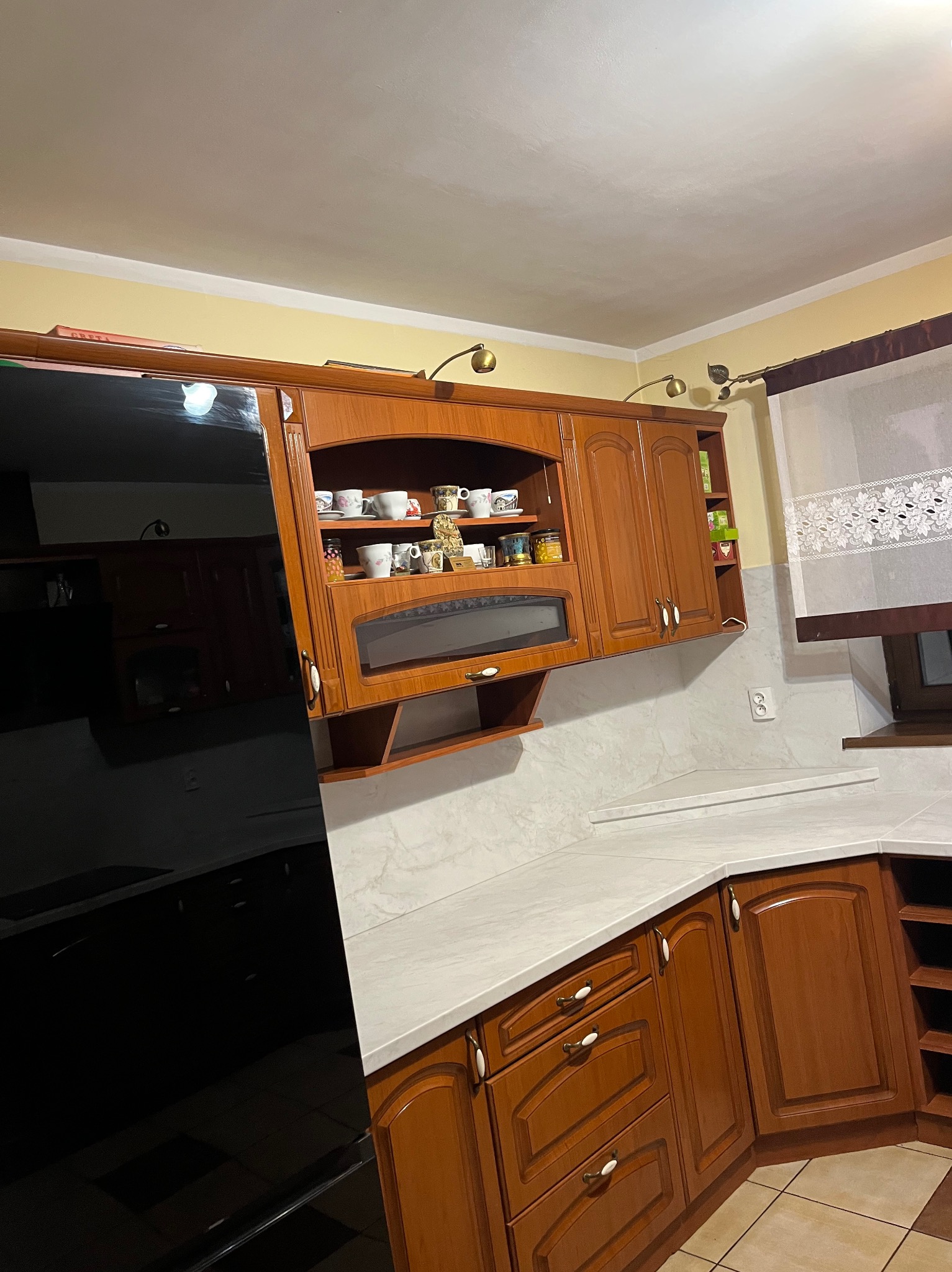 Drewniana zabudowa kuchenna z przeszklonymi szafkami wiszącymi, oświetleniem punktowym i białym blatem. Widoczny fragment czarnej lodówki i okno z firanką.