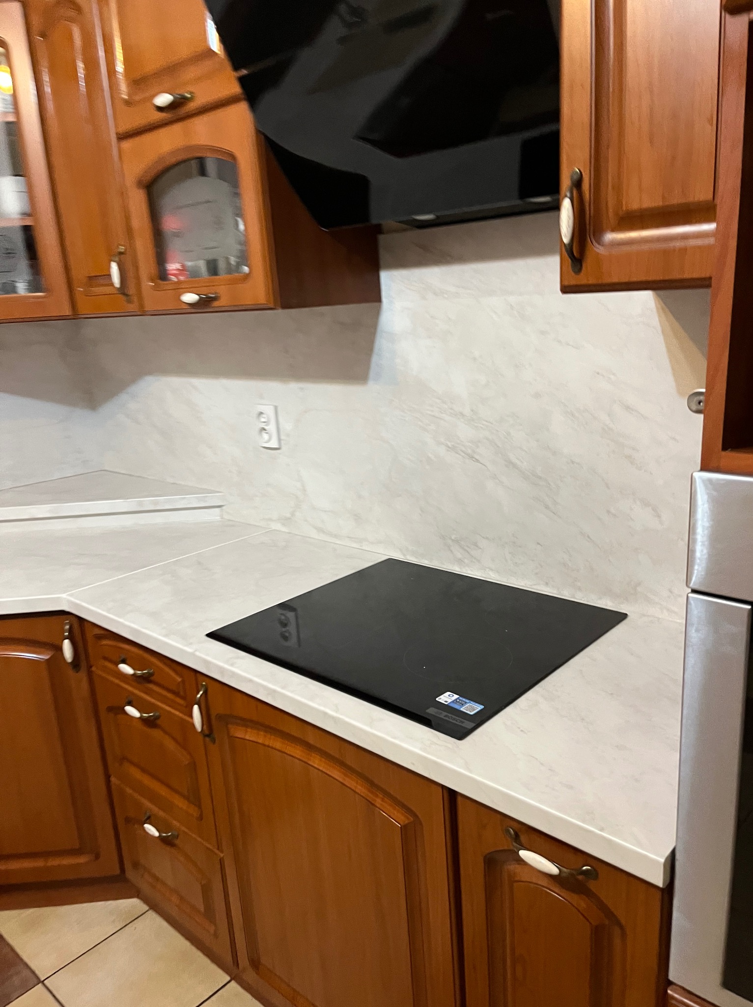 Kuchnia z drewnianymi szafkami, białym blatem i czarną płytą indukcyjną. Widoczny fragment okapu i marmurowa ściana. Klasyczny styl.