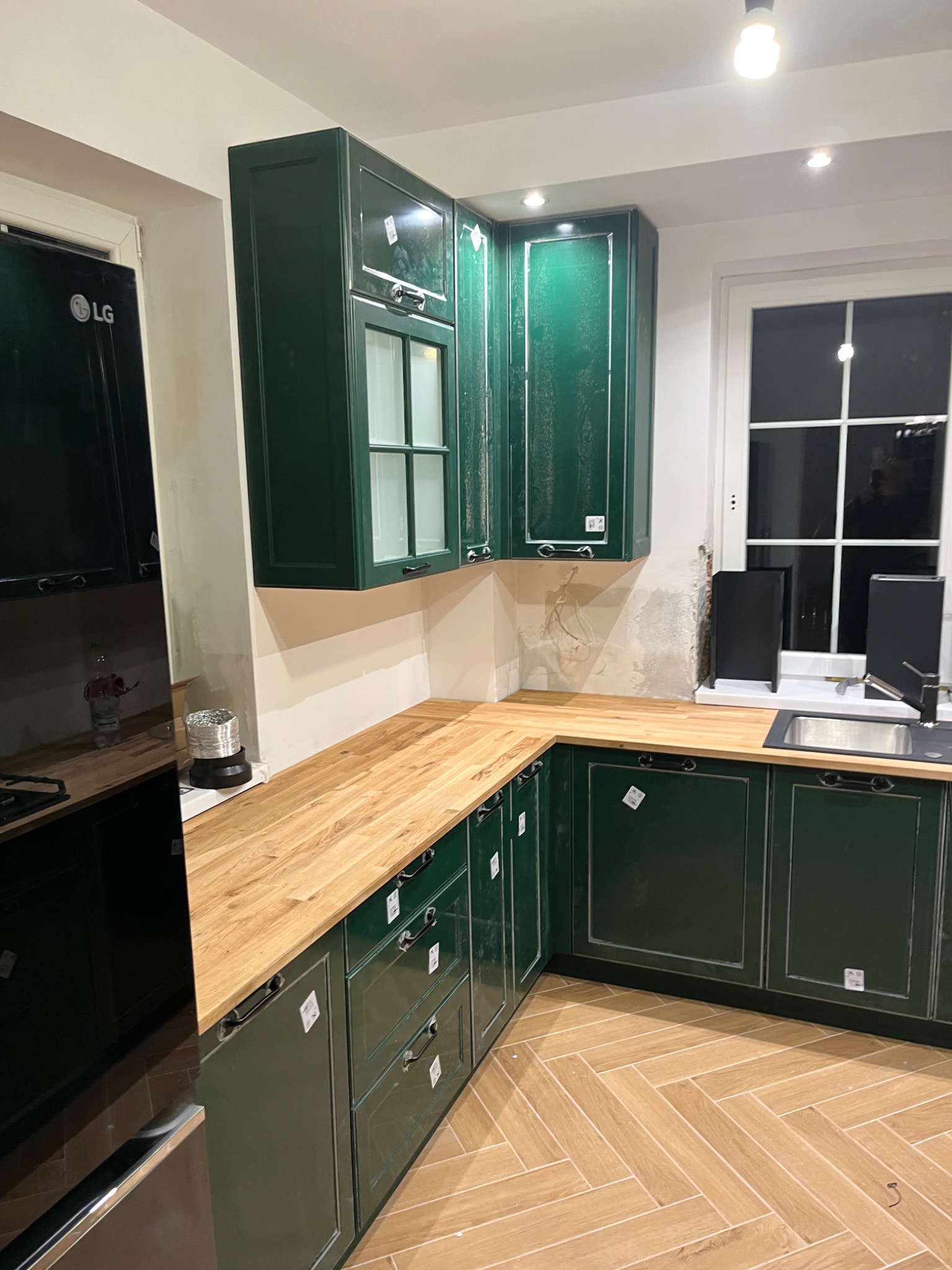 Kuchenne szafki w kolorze butelkowej zieleni z drewnianym blatem, widoczne niedokończone prace wykończeniowe na ścianie i parkiet ułożony w jodełkę.