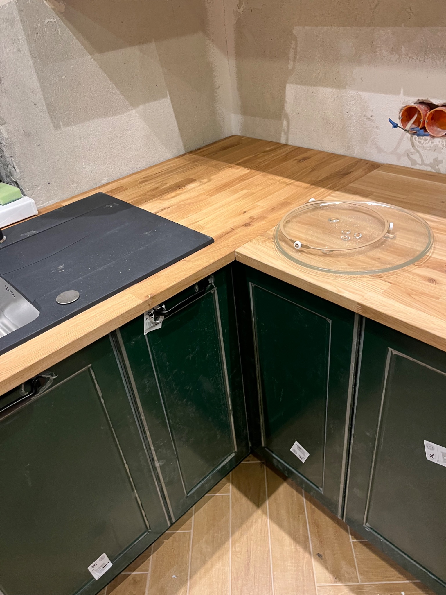 Fragment kuchni w trakcie montażu: drewniany blat, ciemnozielone szafki z frezowanymi frontami i czarny zlewozmywak, widoczne instalacje elektryczne w ścianie i podłoga wyłożona panelami.