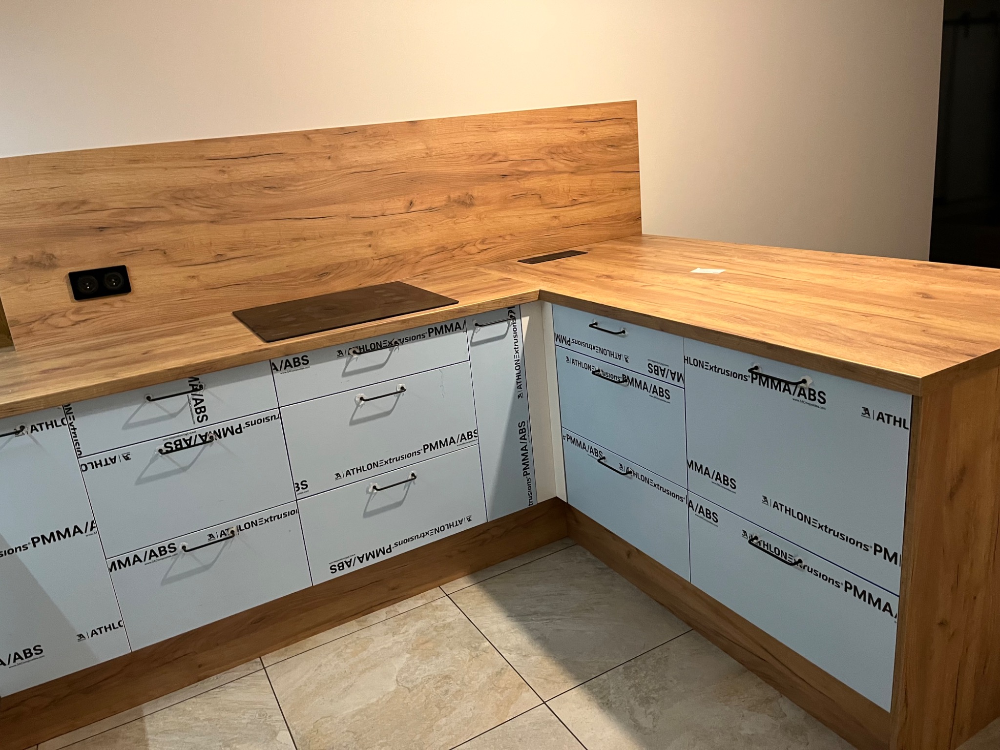 Kuchenna zabudowa narożna z drewnianym blatem i okleiną imitującą drewno, fronty szafek zabezpieczone folią z nadrukiem logo producenta, w blacie zamontowane gniazdka elektryczne i płyta grzewcza.