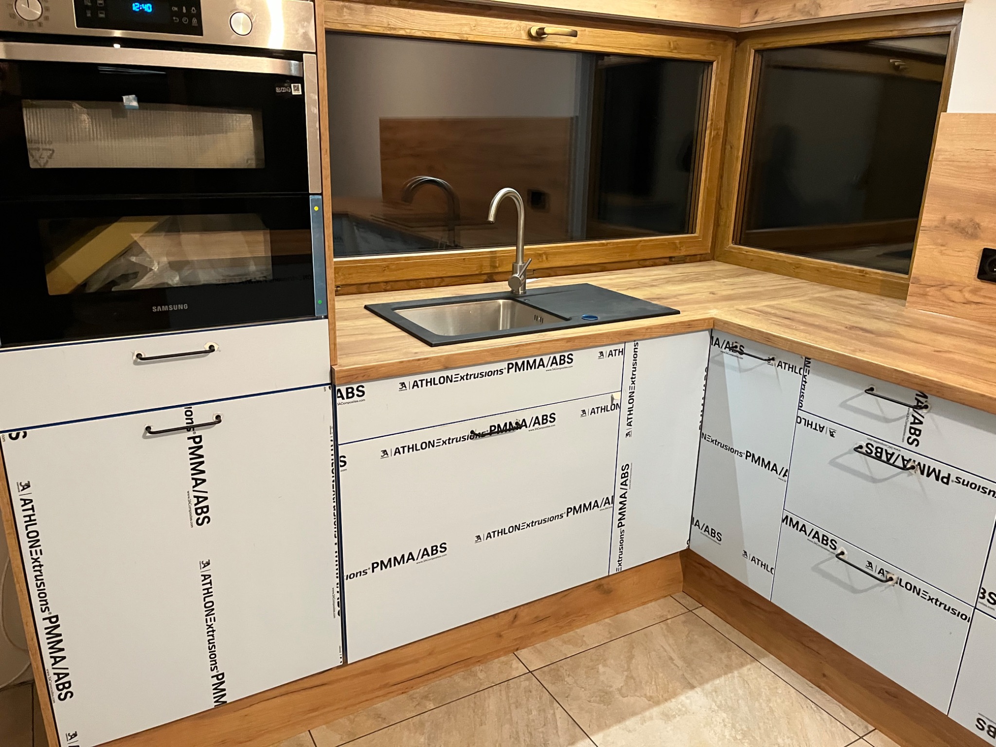 Częściowo zmontowana kuchnia: białe fronty szafek z folią ochronną z nadrukiem, drewniany blat, zlewozmywak ze stalową baterią, okno w drewnianej ramie, piekarnik w zabudowie, widoczne elementy...