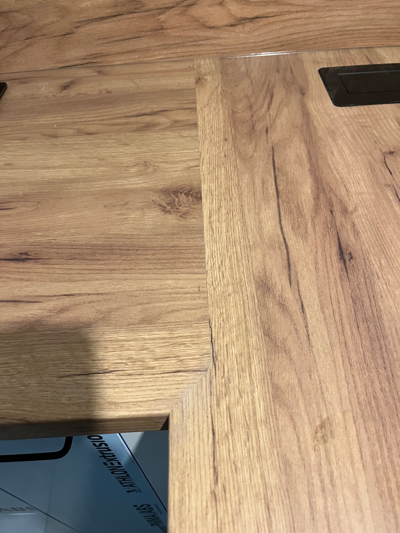 Fragment podłogi z paneli imitujących drewno, z widocznym czarnym gniazdkiem w podłodze i fragmentem blatu meblowego z widocznym napisem 'ATHLON EXTRUSIO'