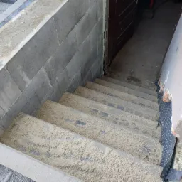 Betonowe schody zewnętrzne w trakcie budowy, z widoczną izolacją fundamentów i zasypanym piaskiem, prowadzące do ciemnego wejścia.