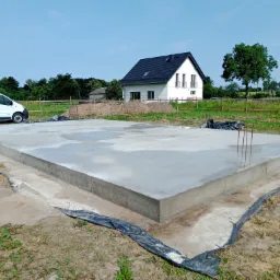 Wylana płyta fundamentowa z wystającymi prętami zbrojeniowymi na tle budowanego domu jednorodzinnego i zaparkowanego białego busa.