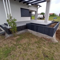 Firma Budowlana MatBud - Nowoczesny taras z szarych płyt, czarne obrzeża rabatki, białe słupy podtrzymujące zadaszenie, widok na ogród i zaparkowany biały samochód dostawczy.