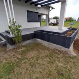 Nowoczesny taras z szarych płyt, czarne obrzeża rabatki, białe słupy podtrzymujące zadaszenie, widok na ogród i zaparkowany biały samochód dostawczy.