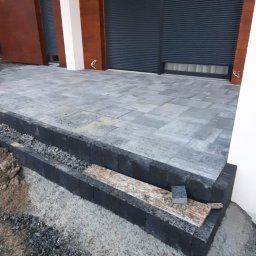 Firma Budowlana MatBud - Wykończony taras z szarej kostki brukowej i czarnych obrzeży, widoczne fragmenty elewacji budynku z brązowymi panelami i szarymi roletami zewnętrznymi.