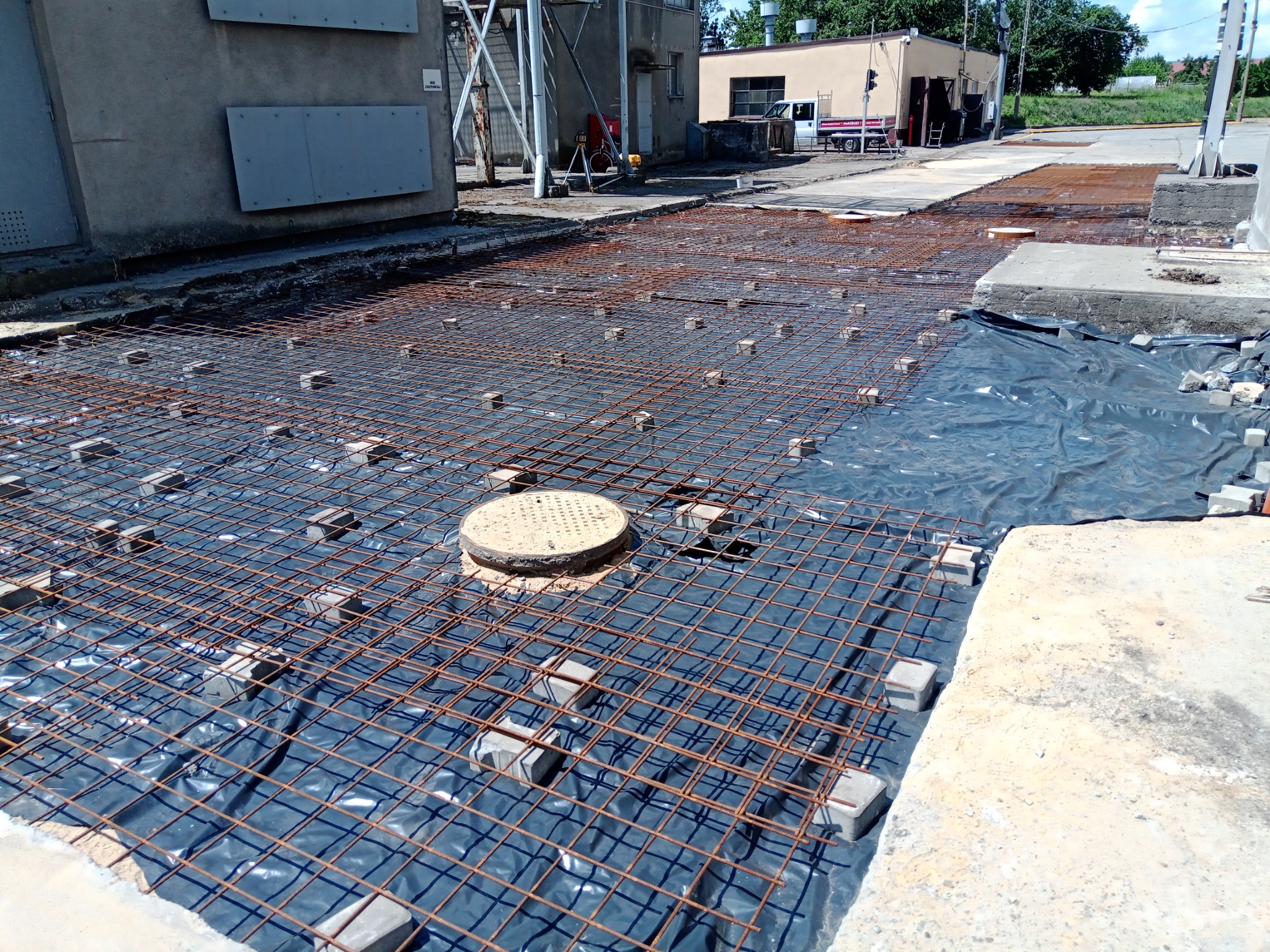 Przygotowanie terenu pod wylewkę betonową: siatka zbrojeniowa rozłożona na czarnej folii, podparta betonowymi kostkami, widoczny właz kanalizacyjny i fragmenty istniejącej betonowej nawierzchni.