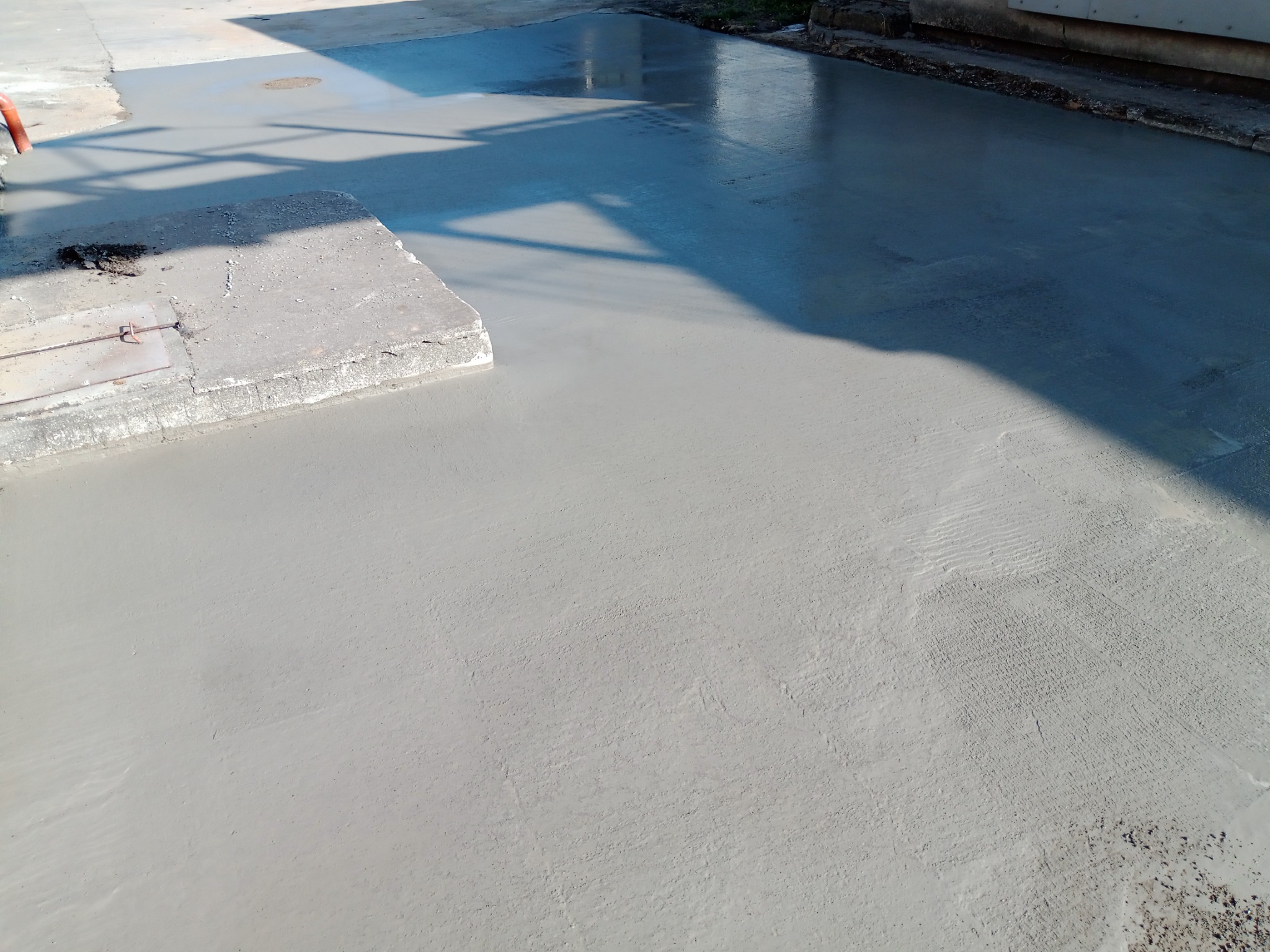 Świeżo wylana, gładka wylewka betonowa na zewnątrz budynku, częściowo mokra, z widocznym włazem kanalizacyjnym i cieniem