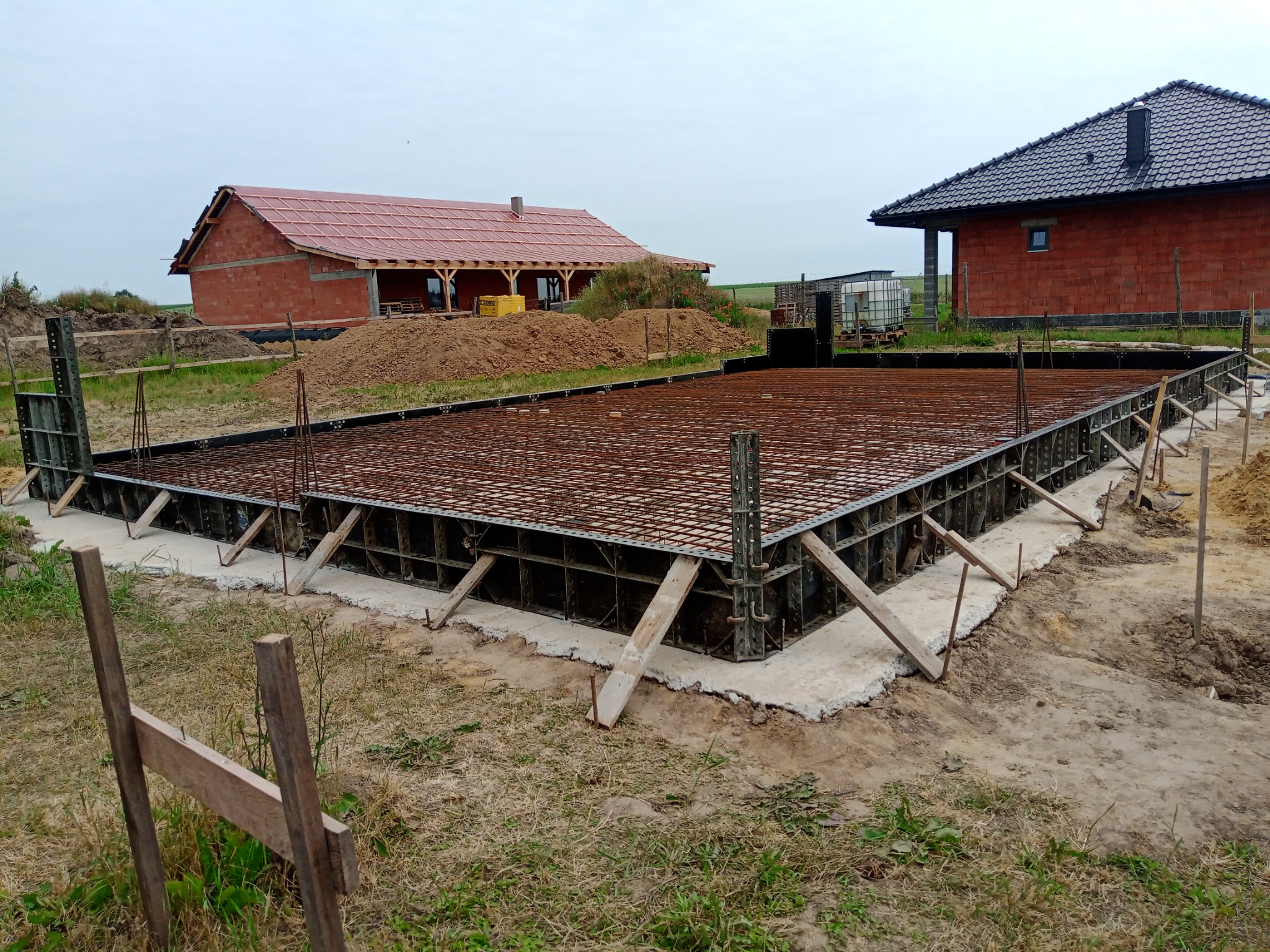 Zbrojenie fundamentu w szalunku stalowym, widoczne kotwy i drewniane podpory, w tle budynki w stanie surowym otwarte.