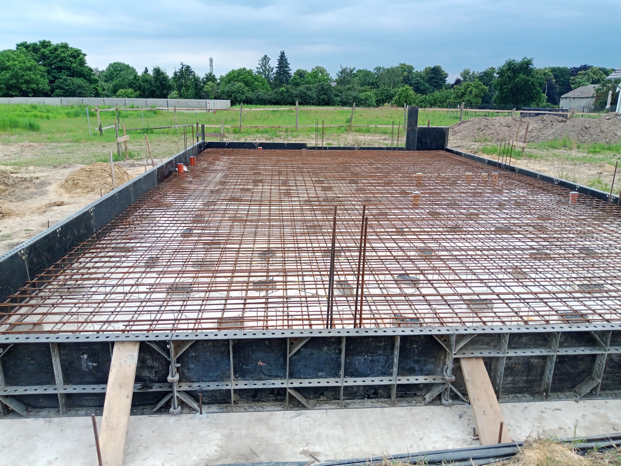 Zbrojenie płyty fundamentowej z widocznym szalunkiem i folią izolacyjną, przygotowane do zalania betonem na tle zieleni i pochmurnego nieba.