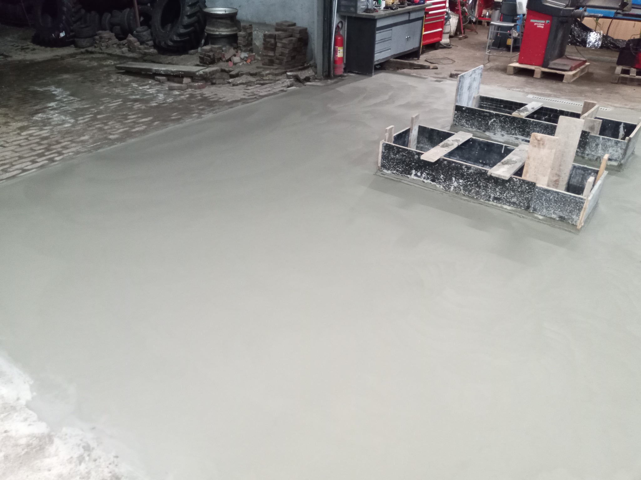 Świeżo wylana, gładka betonowa posadzka w hali warsztatowej, z widocznymi drewnianymi formami szalunkowymi i oponami w tle.