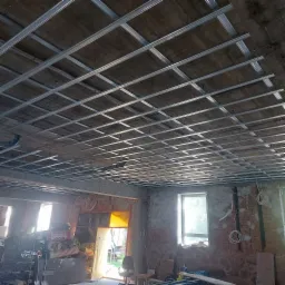 Metalowa konstrukcja sufitowa z profili w surowym wnętrzu, widoczne okna i materiały budowlane w tle, etap przed montażem płyt.